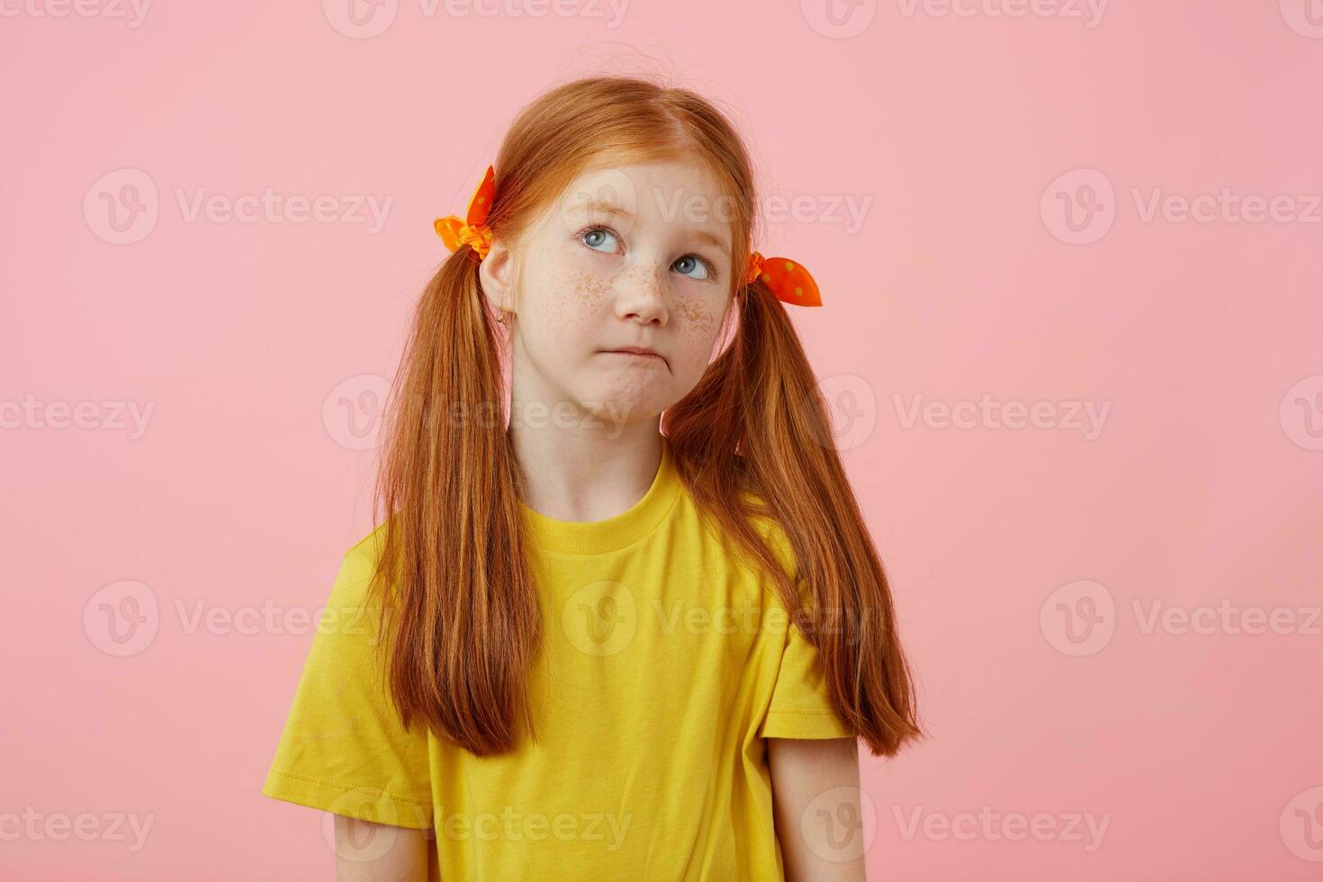 ritratto poco pensiero lentiggini dai capelli rossi ragazza con Due code, sembra lontano, tocchi guance, indossa nel giallo maglietta, sta al di sopra di rosa sfondo con copia spazio. foto