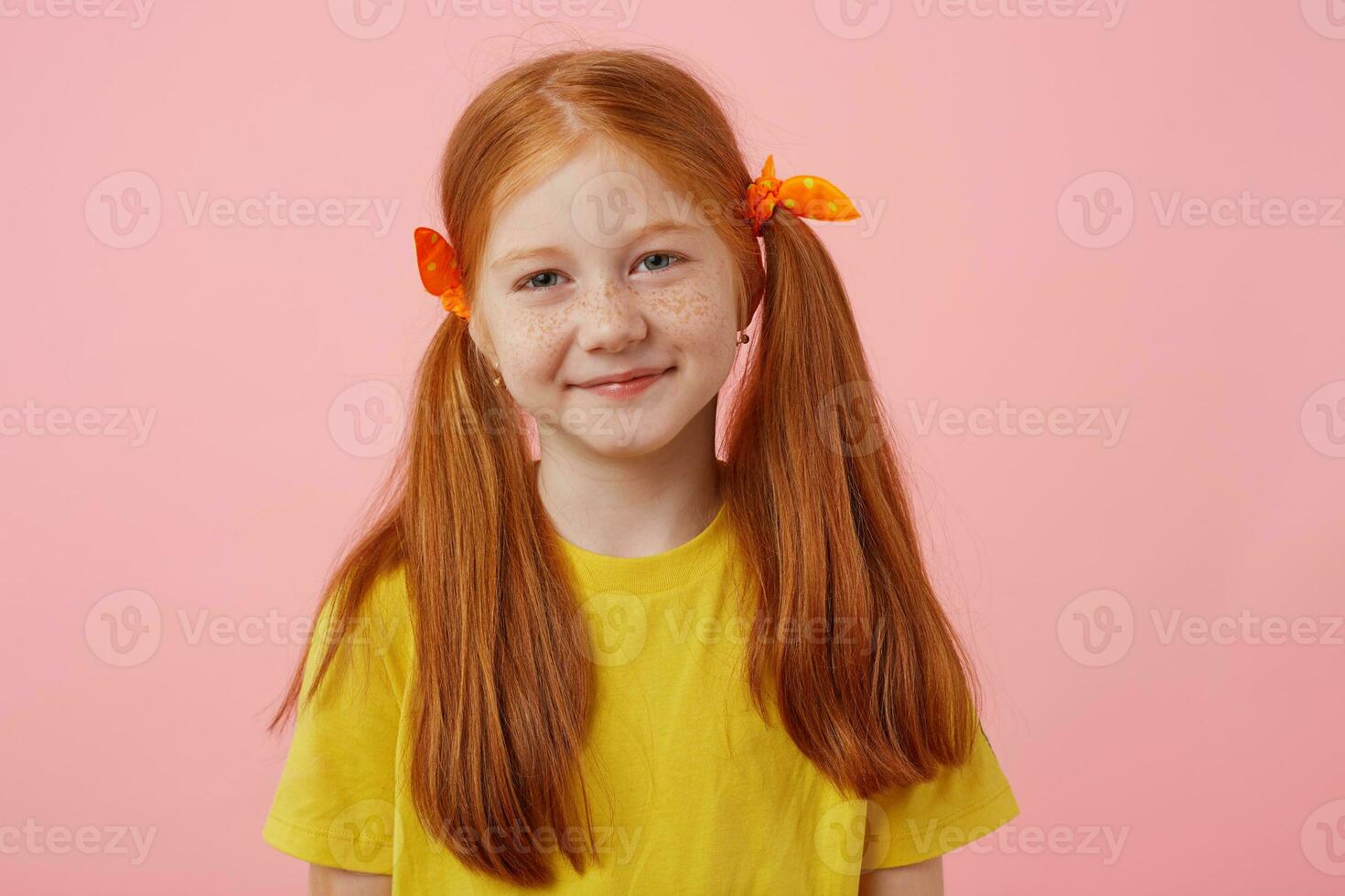 vicino su di contento petite lentiggini dai capelli rossi ragazza con Due code, sorridente e sembra carino, indossa nel giallo maglietta, sta al di sopra di rosa sfondo. foto