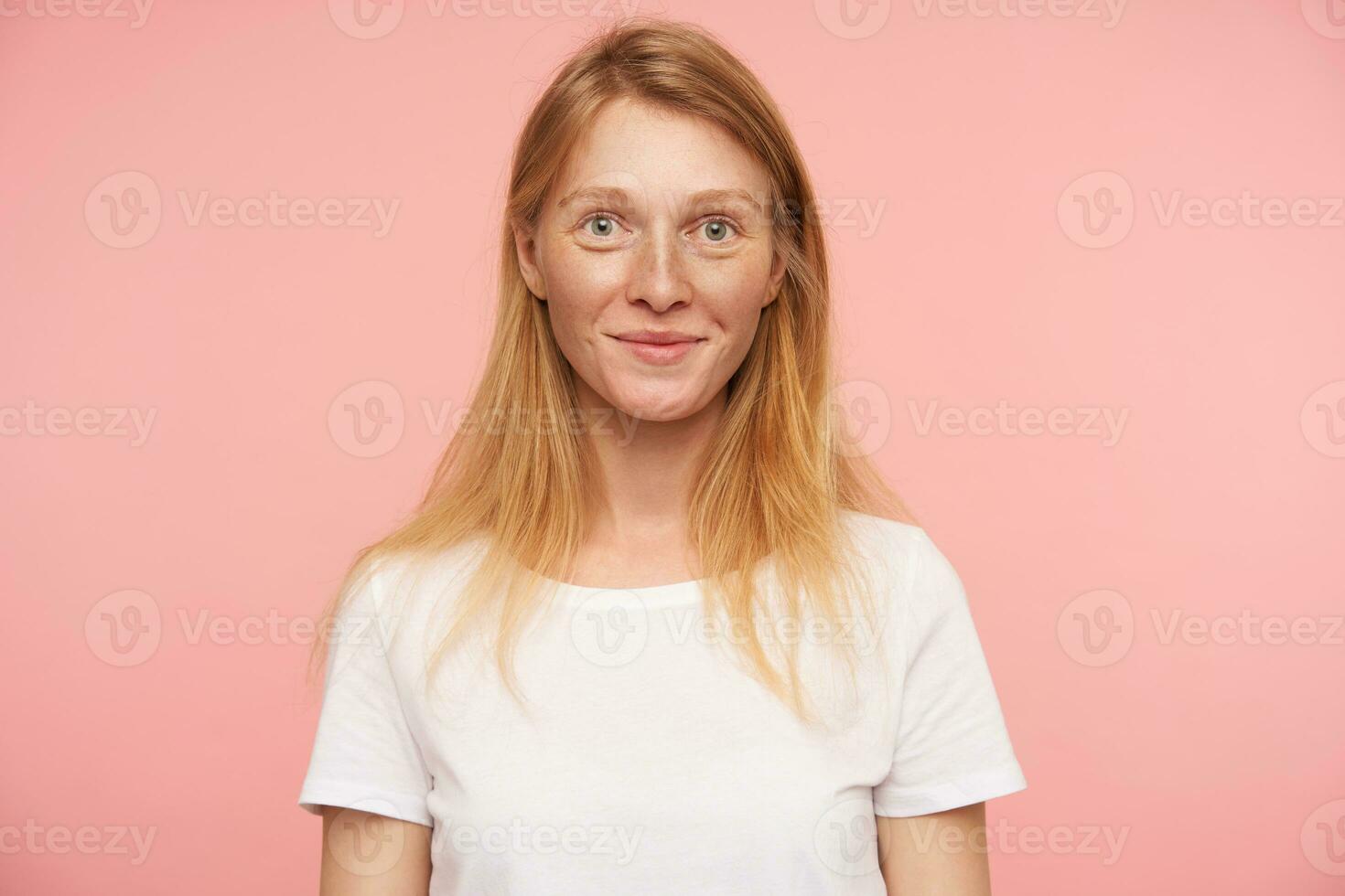 ritratto di affascinante giovane testa Rossa signora con naturale trucco guardare positivamente a telecamera e sorridente piacevolmente, in posa al di sopra di rosa sfondo con mani giù foto