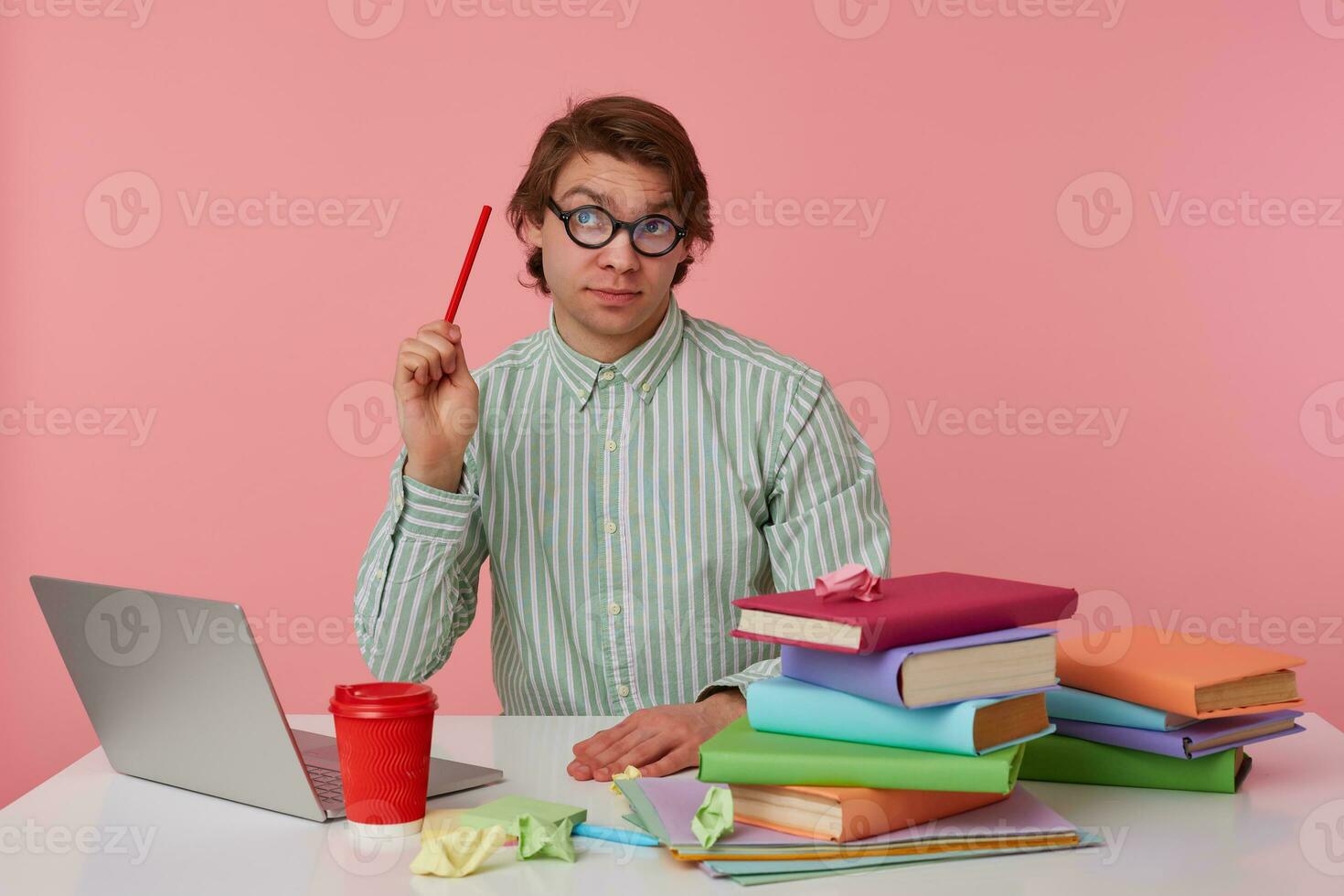 pensiero giovane uomo nel bicchieri si siede di il tavolo e Lavorando con computer portatile, sembra su, detiene nel mano un' matita, provando per risolvere un' difficile equazione, isolato al di sopra di rosa sfondo. foto