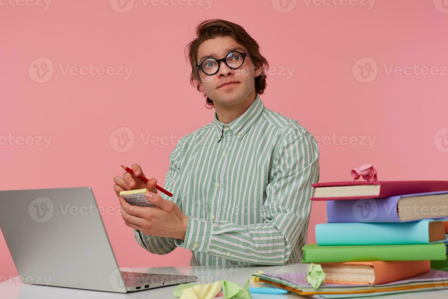 giovane pensiero uomo nel bicchieri si siede di il tavolo e Lavorando con computer portatile, detiene nel mano un' matita, accigliato e scrive il idea su il etichetta, sembra su isolato al di sopra di rosa sfondo. foto