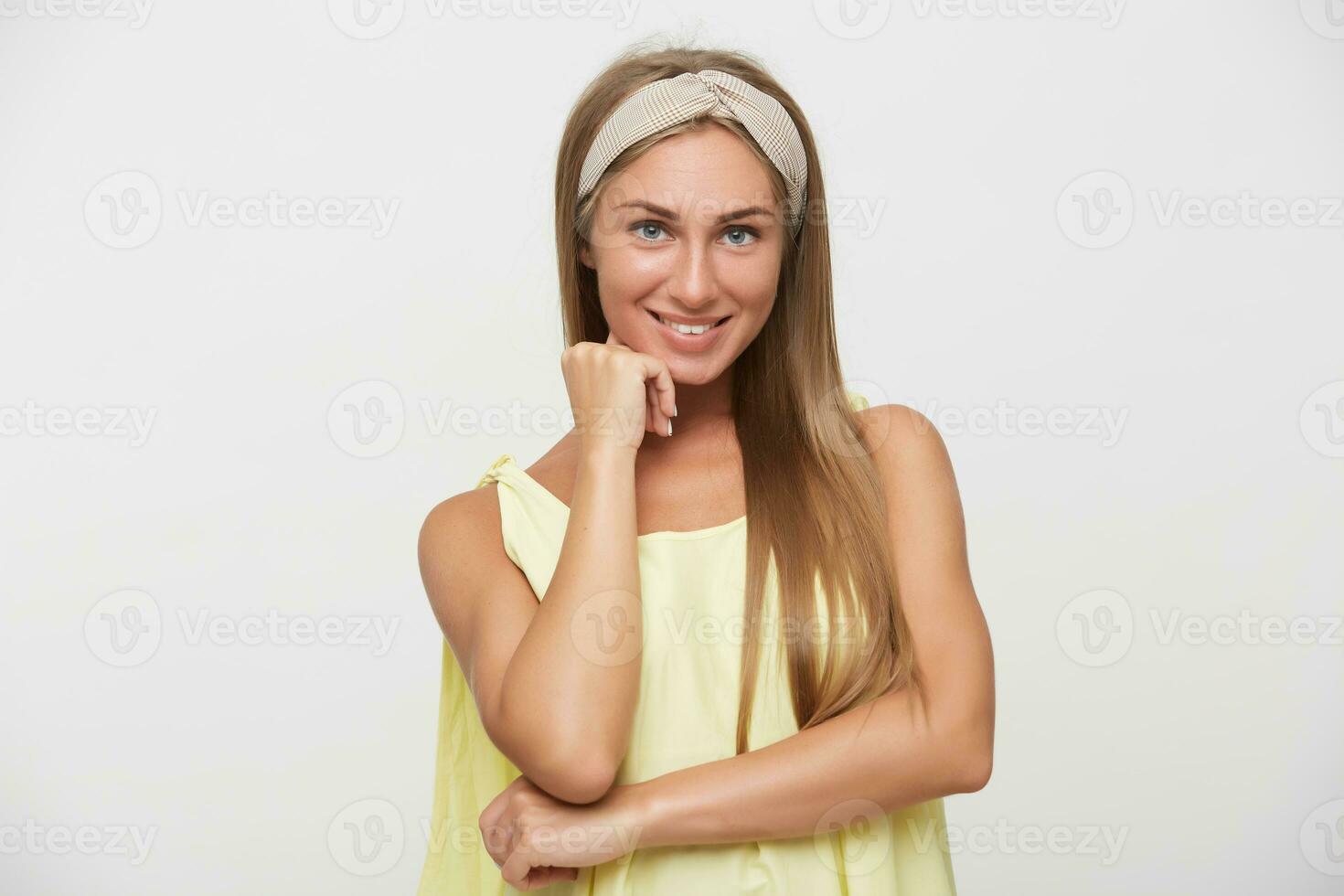 ritratto di giovane bellissimo allegro lungo dai capelli bionda signora indossare beige fascia per capelli mentre in posa al di sopra di bianca sfondo, sorridente piacevolmente per telecamera e Tenere sollevato mano su sua viso foto