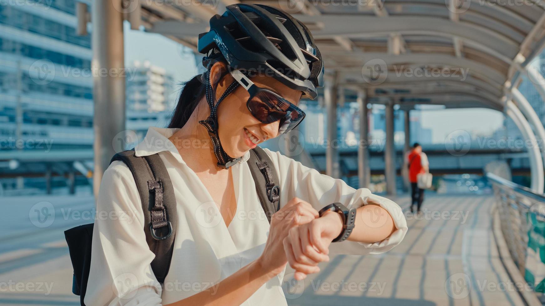 donna d'affari asiatica con bicicletta zaino sorridente guardare smartwatch in strada di città andare a lavorare in ufficio. la ragazza sportiva usa la sua app per l'orologio per il monitoraggio del fitness. andare al lavoro, pendolare d'affari in città. foto