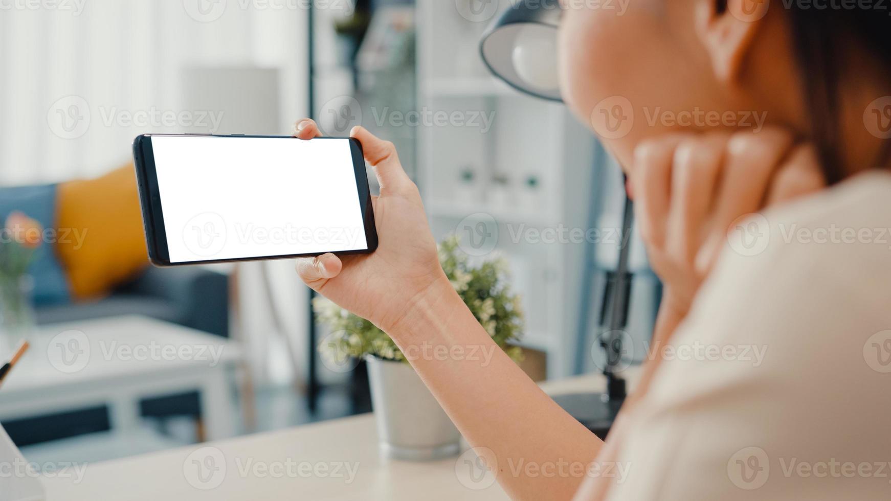 la giovane donna asiatica usa lo smartphone con lo schermo bianco vuoto finto display per il testo pubblicitario mentre lavora in modo intelligente da casa in soggiorno. tecnologia chiave di crominanza, concetto di design di marketing. foto