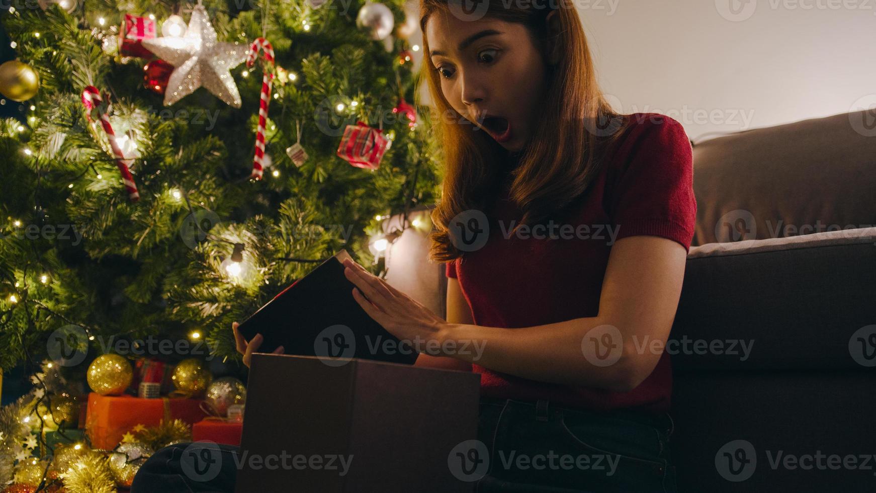 giovane donna asiatica che si diverte ad aprire la scatola regalo di Natale vicino all'albero di Natale decorato con ornamenti nel soggiorno di casa. buona notte di natale e felice anno nuovo festa delle vacanze. foto