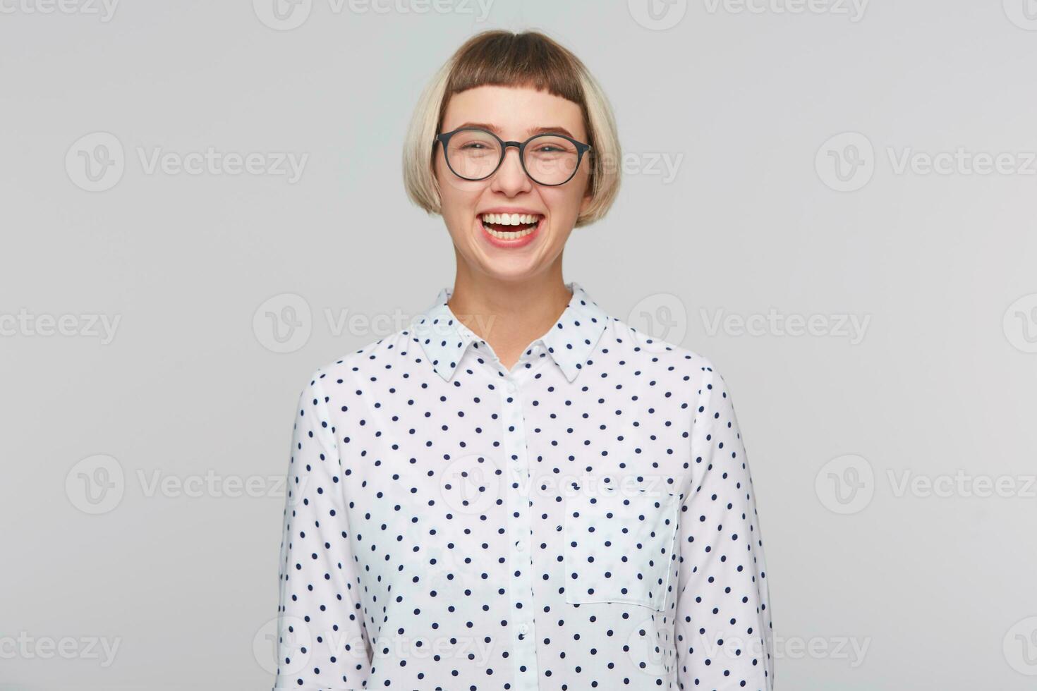 ritratto di allegro bella bionda giovane donna indossa polka punto camicia e bicchieri si sente contento e ridendo isolato al di sopra di bianca sfondo foto