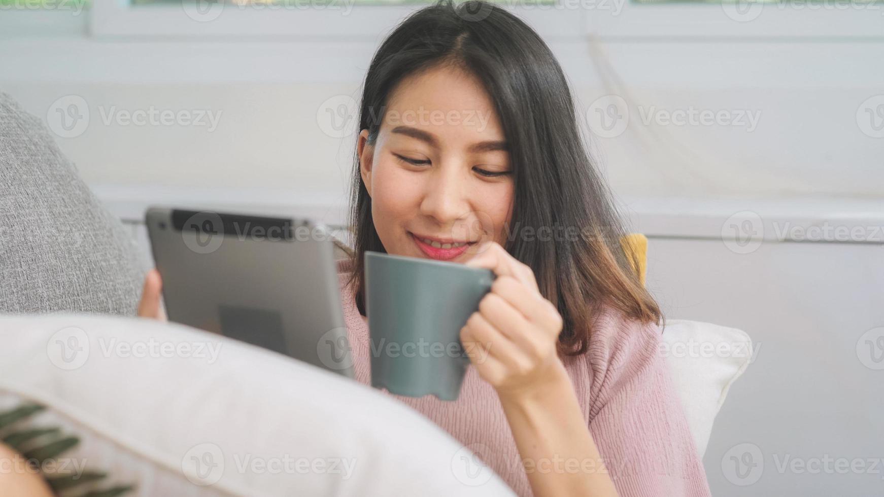 bella donna asiatica sorridente attraente che utilizza compressa che tiene una tazza calda di caffè o tè mentre è sdraiata sul divano quando si rilassa nel soggiorno di casa. donne di stile di vita a casa concetto. foto