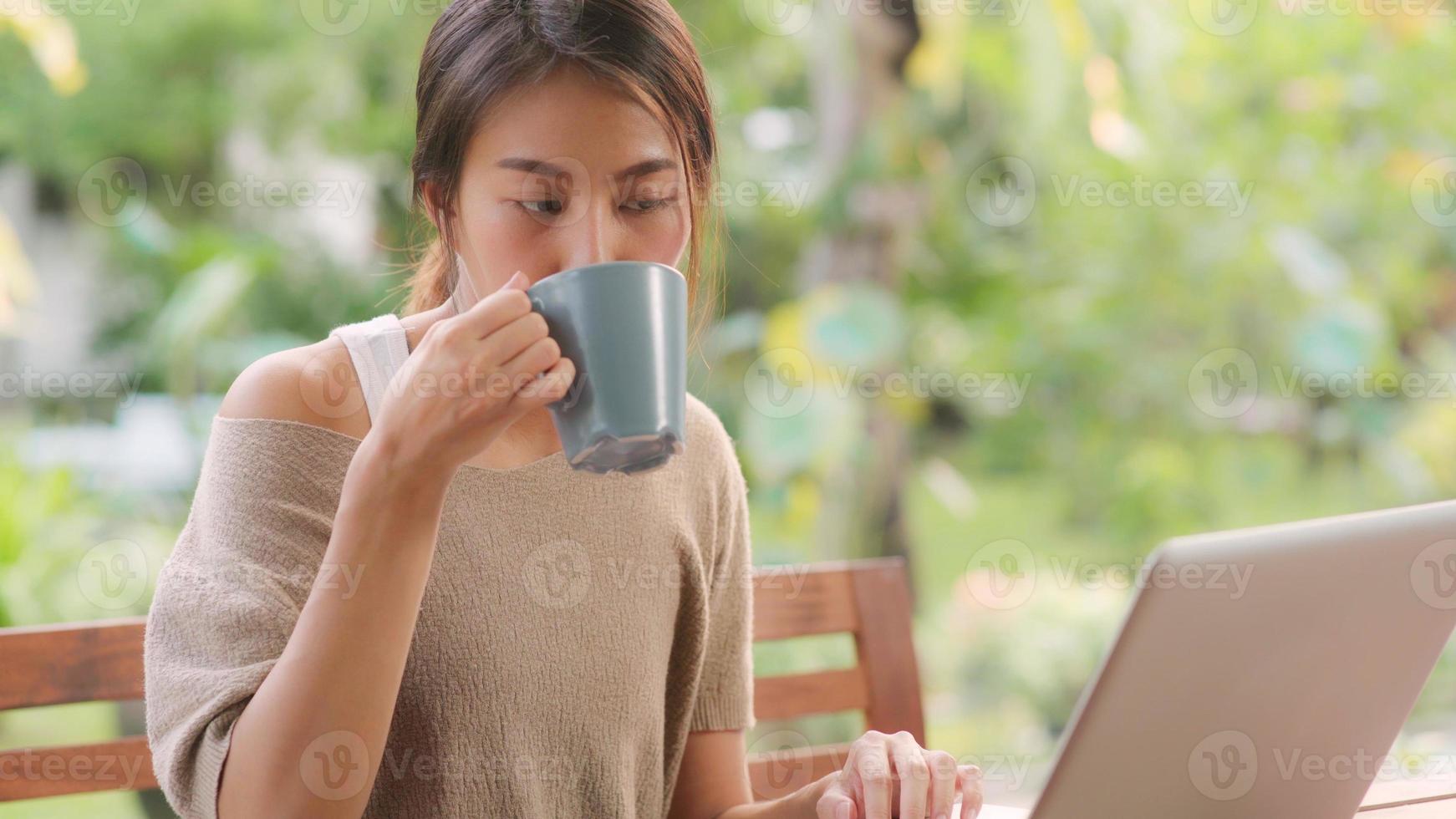donna asiatica freelance che lavora a casa, donna d'affari che lavora al computer portatile e beve caffè seduta sul tavolo in giardino al mattino. donne di stile di vita che lavorano a casa concetto. foto