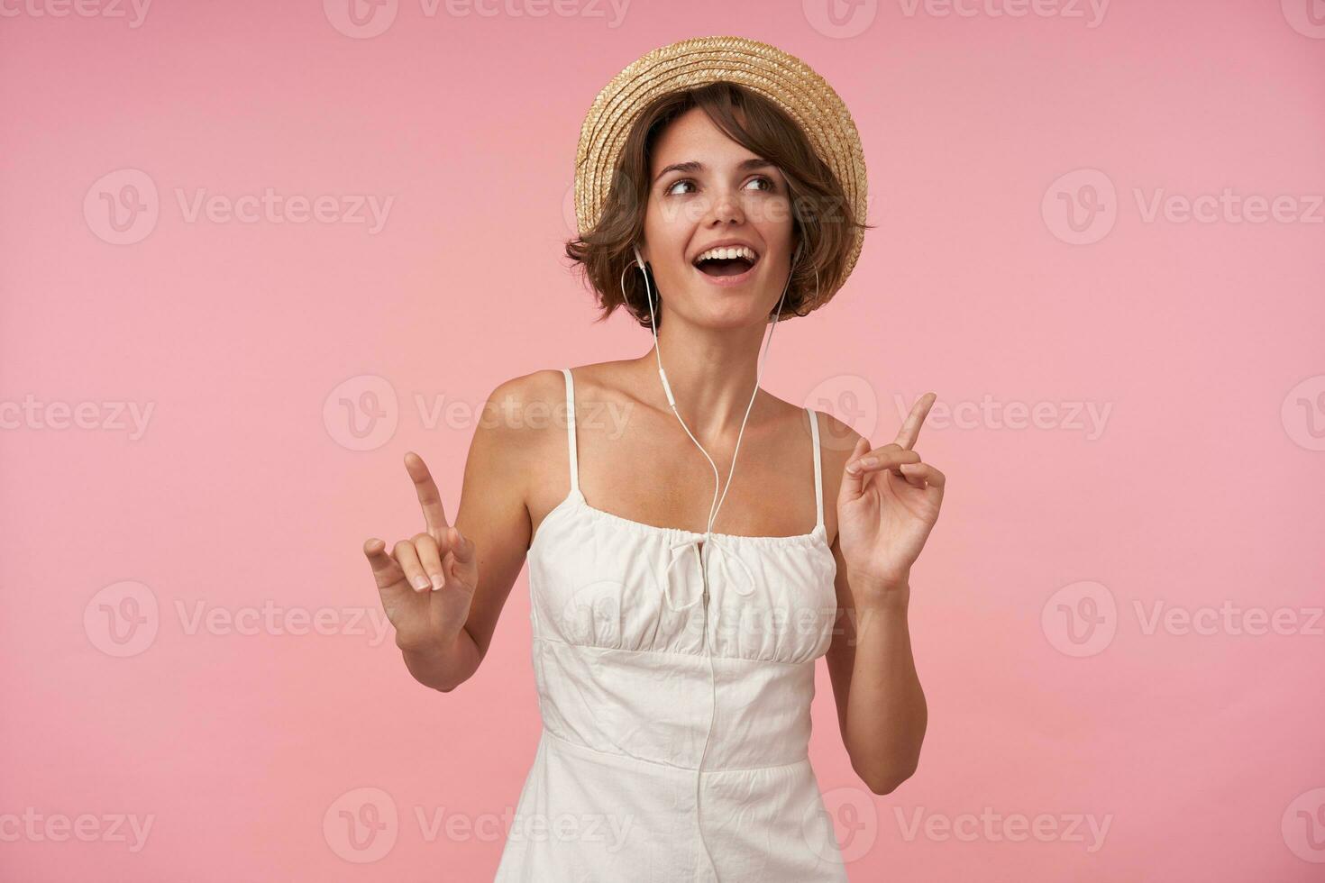 attraente giovane brunetta femmina con casuale acconciatura raccolta mani mentre ascoltando per bene musica, guardare a parte allegramente mentre in piedi al di sopra di rosa sfondo foto