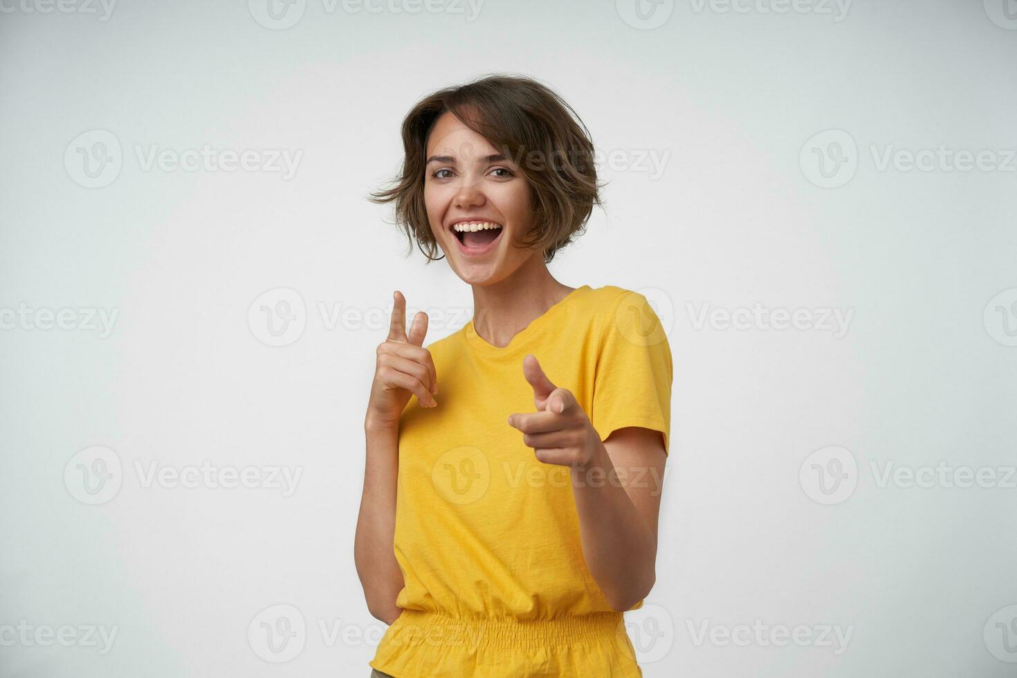 contento bellissimo giovane signora con casuale acconciatura puntamento avanti con sollevato indici, guardare a telecamera con gioia e sorridente ampiamente, in piedi al di sopra di bianca sfondo nel giallo maglietta foto