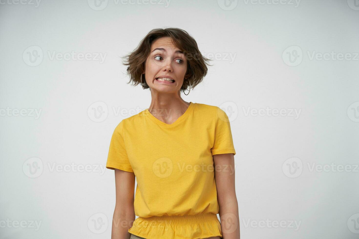 ritratto di imbarazzato giovane femmina signora con corto Marrone capelli guardare a parte e mostrando denti confusamente, conservazione mani lungo sua corpo mentre in posa al di sopra di bianca sfondo foto