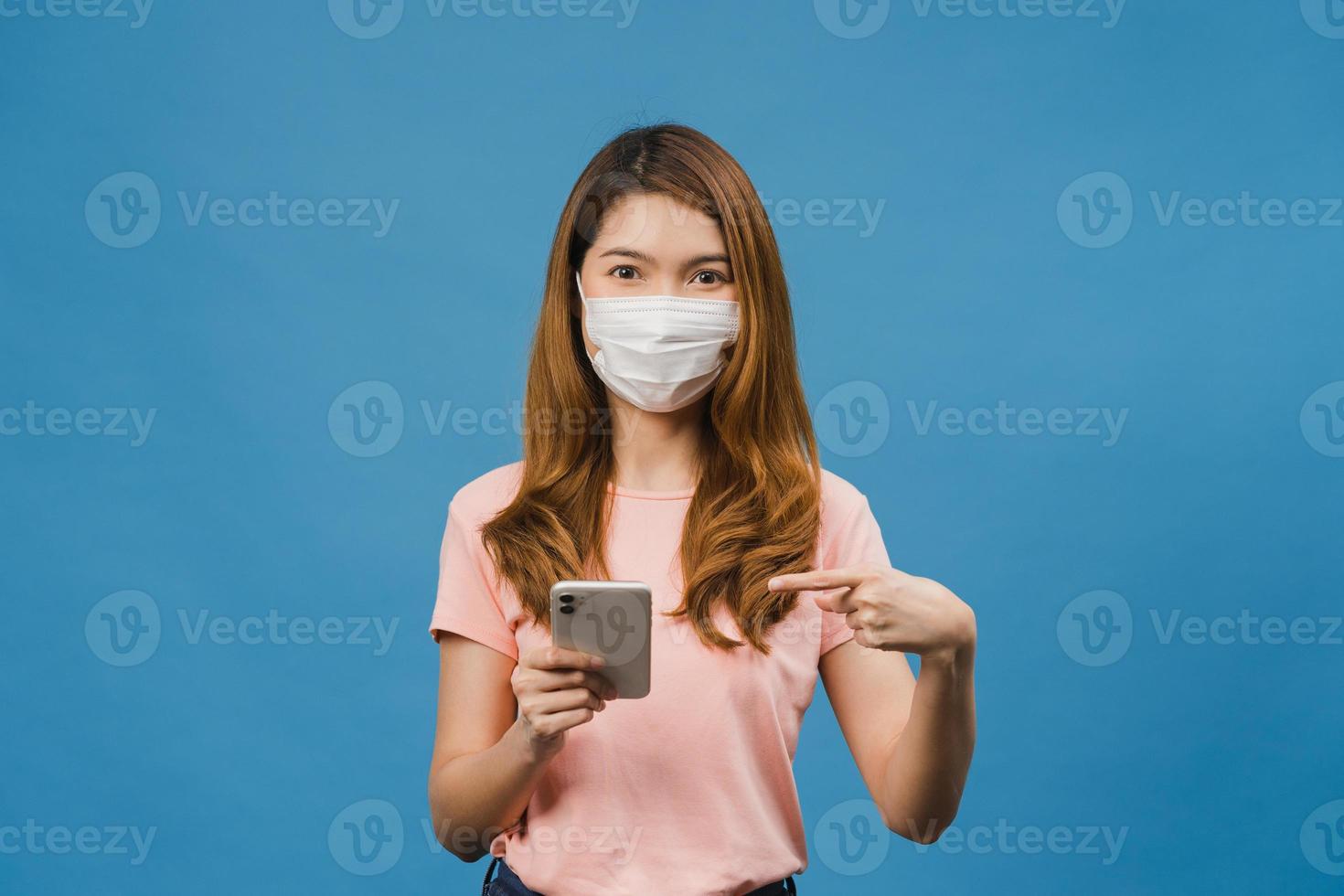 giovane ragazza asiatica che indossa una maschera medica utilizzando il telefono cellulare con vestiti in abiti casual isolati su sfondo blu. autoisolamento, distanziamento sociale, quarantena per la prevenzione del virus corona. foto