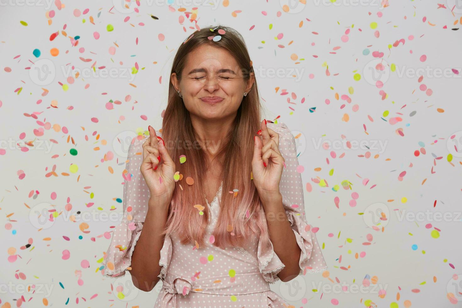 sorridente carino giovane donna con attraversato dita e chiuso occhi indossa polka punto rosa vestito festeggiare compleanno e fa un' desiderio isolato al di sopra di bianca sfondo con coriandoli foto