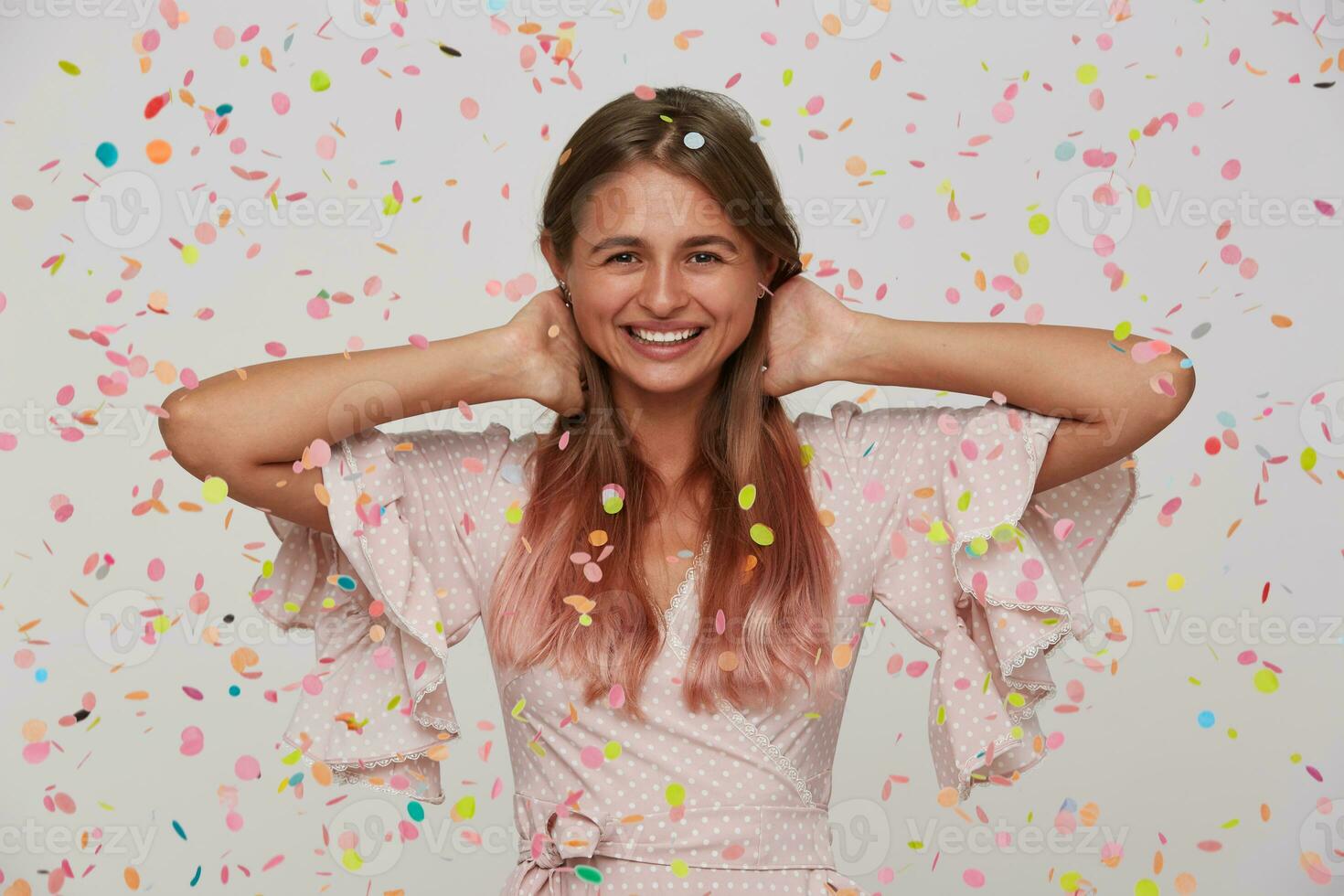 avvicinamento di contento bella giovane donna con lungo tinti pastello rosa capelli indossa polka punto rosa vestito festeggiare compleanno e avendo divertimento al di sopra di bianca sfondo con coriandoli foto