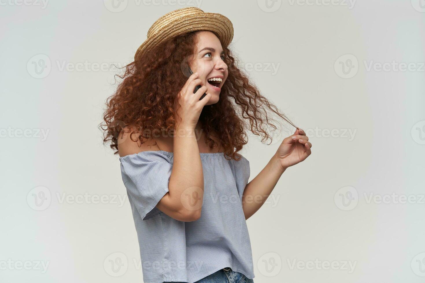 simpatico guardare donna, bellissimo ragazza con Riccio Zenzero capelli. indossare a strisce spalle scoperte camicetta e cappello. parlando su un' smartphone. parlare con amico su un' Telefono. In piedi isolato al di sopra di bianca sfondo foto