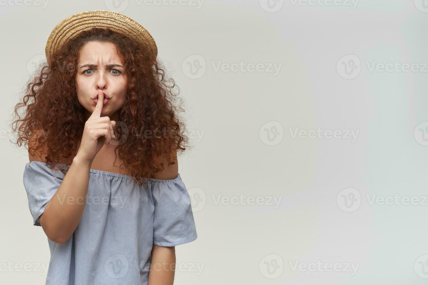 adolescenziale ragazza, donna con Riccio Zenzero capelli. indossare a strisce spalle scoperte camicetta e cappello. mostrando silenzio cartello, cipiglio. Guardando a il telecamera, isolato al di sopra di bianca sfondo. copia spazio su il giusto foto