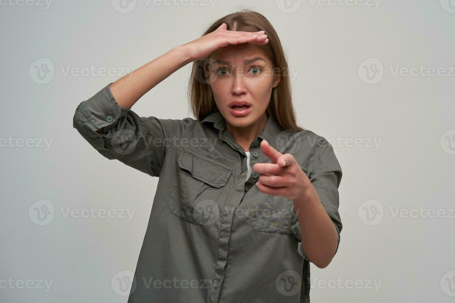 giovane signora, bella donna con Marrone lungo capelli. indossare grigio camicia, detiene sua mano al di sopra di sua fronte, puntamento a voi. Guardando con preoccupato viso a il telecamera isolato al di sopra di grigio sfondo foto