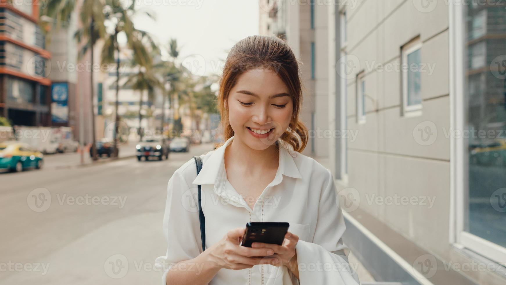 giovane imprenditrice asiatica di successo in abiti da ufficio di moda utilizzando lo smartphone e digitando un messaggio di testo mentre si cammina da soli all'aperto nella città moderna urbana al mattino. concetto di affari in movimento. foto