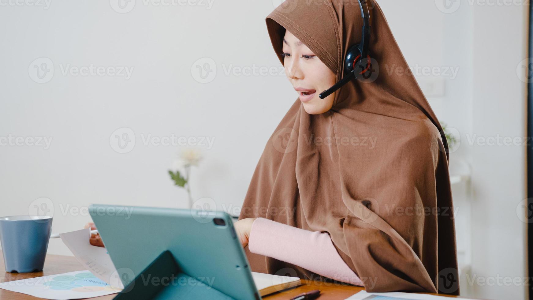 la signora musulmana asiatica indossa le cuffie utilizzando il tablet digitale parla con i colleghi del rapporto di vendita nella videochiamata in conferenza mentre si lavora da casa in cucina. distanziamento sociale, quarantena per il virus corona. foto