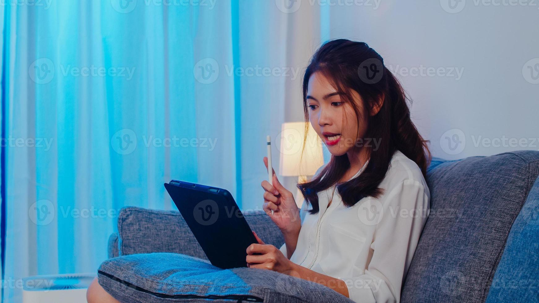 donna d'affari asiatica che utilizza tablet parla con i colleghi del piano in videochiamata mentre lavora da casa in soggiorno di notte. autoisolamento, distanziamento sociale, quarantena per la prevenzione del coronavirus. foto