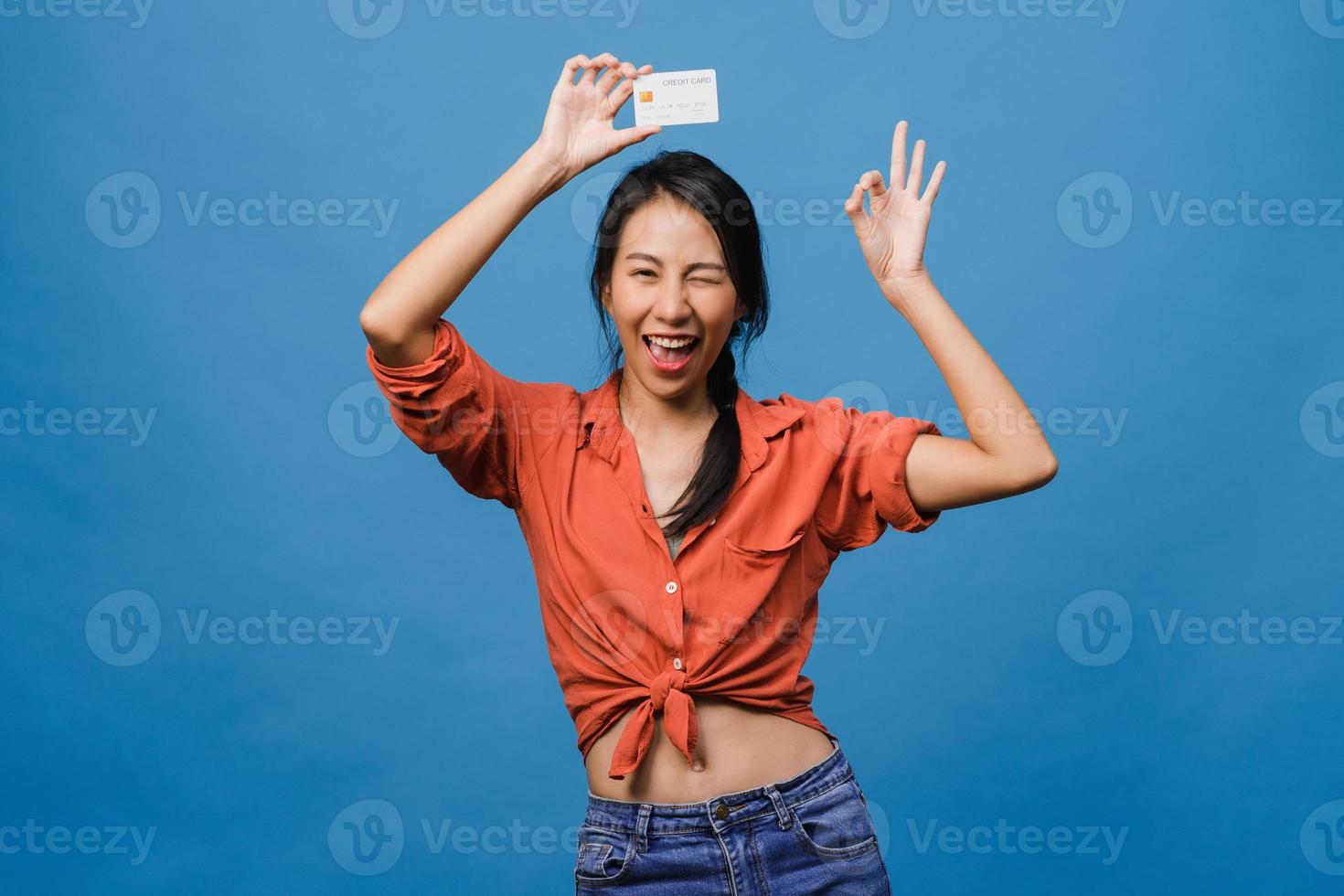 la giovane donna asiatica mostra la carta di credito con l'espressione positiva, sorride ampiamente, vestita con abiti casual sentendo felicità e stando isolata su sfondo blu. concetto di espressione facciale. foto