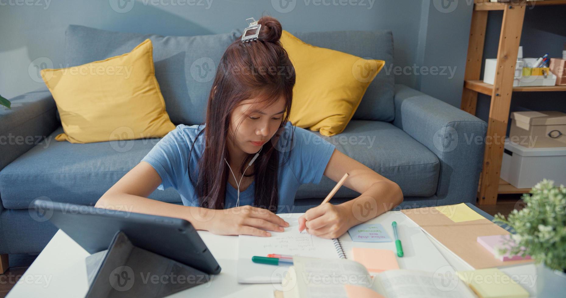 giovane ragazza asiatica adolescente con cuffie per abbigliamento casual usa tablet digitale impara taccuino di lettura online di scrittura nel soggiorno di casa. isolare l'istruzione online e-learning concetto di pandemia di coronavirus. foto