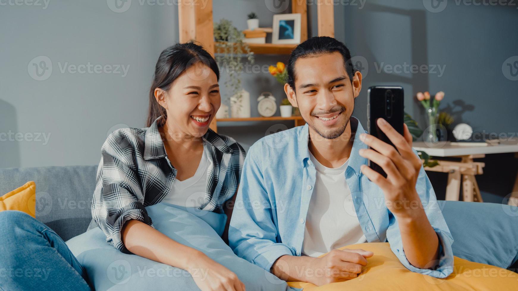 felice giovane coppia asiatica uomo e donna sedersi sul divano usa la videochiamata facetime dello smartphone con gli amici e la famiglia nel soggiorno di casa. resta a casa quarantena, distanza sociale, concetto di giovane sposato. foto