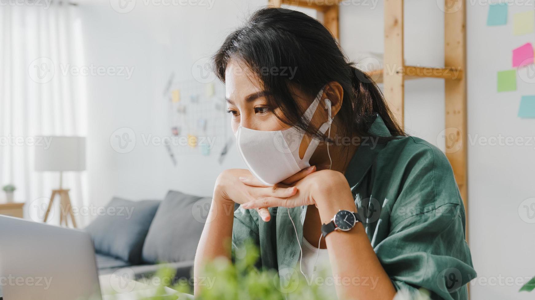 donna d'affari asiatica che indossa una maschera medica utilizzando il laptop parla con i colleghi del piano in videochiamata mentre lavora da casa in soggiorno. distanziamento sociale, quarantena per la prevenzione del virus corona. foto