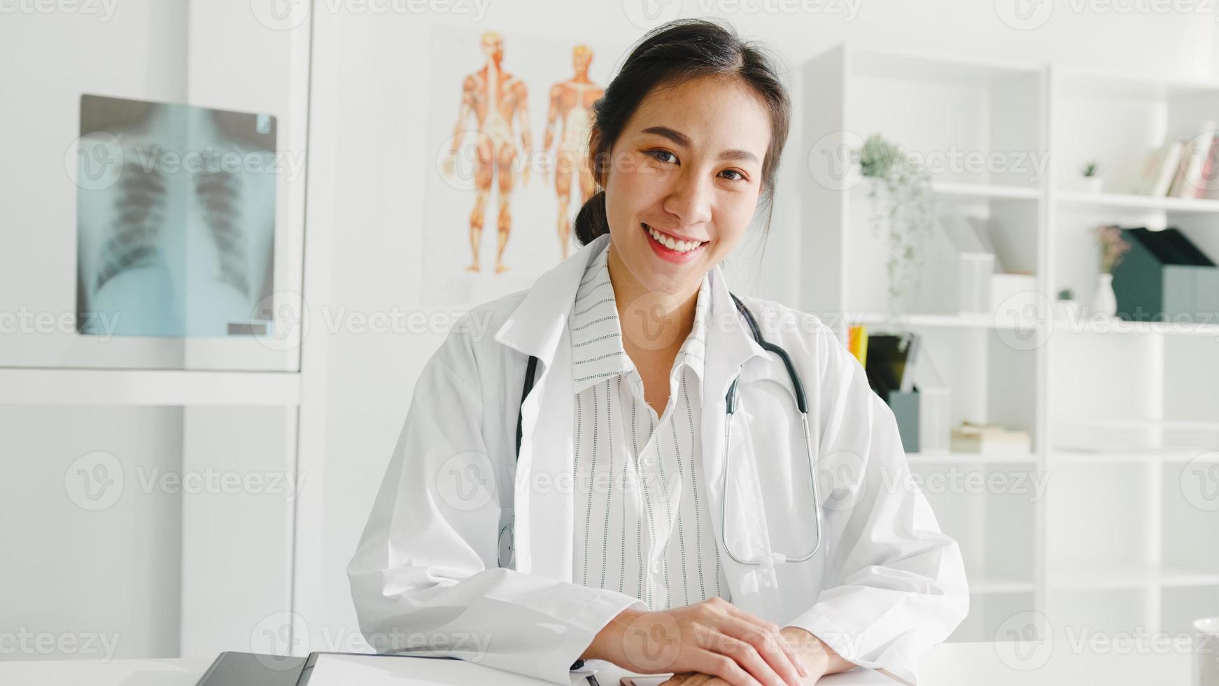 fiducioso giovane dottoressa asiatica in uniforme medica bianca con stetoscopio che guarda l'obbiettivo e sorride durante la videoconferenza con il paziente in ospedale. concetto di consulenza e terapia. foto