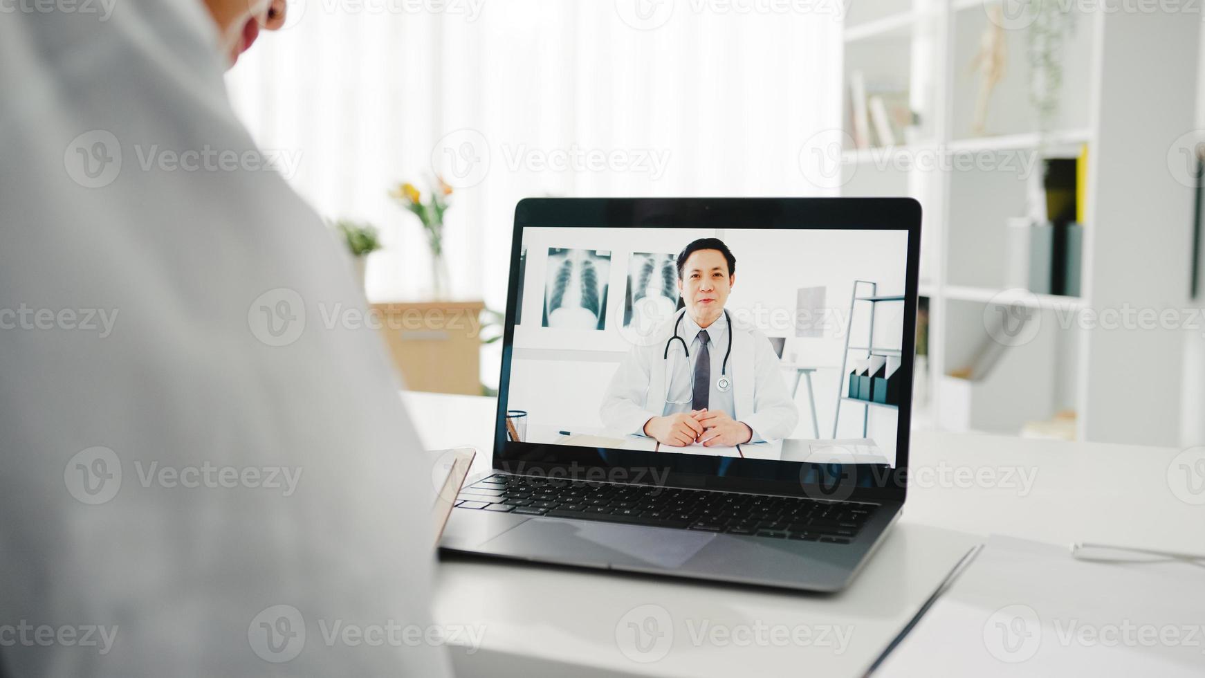 giovane dottoressa asiatica in uniforme medica bianca utilizzando laptop parlando in videoconferenza con un medico anziano alla scrivania in clinica o ospedale. distanziamento sociale, quarantena per il virus corona. foto