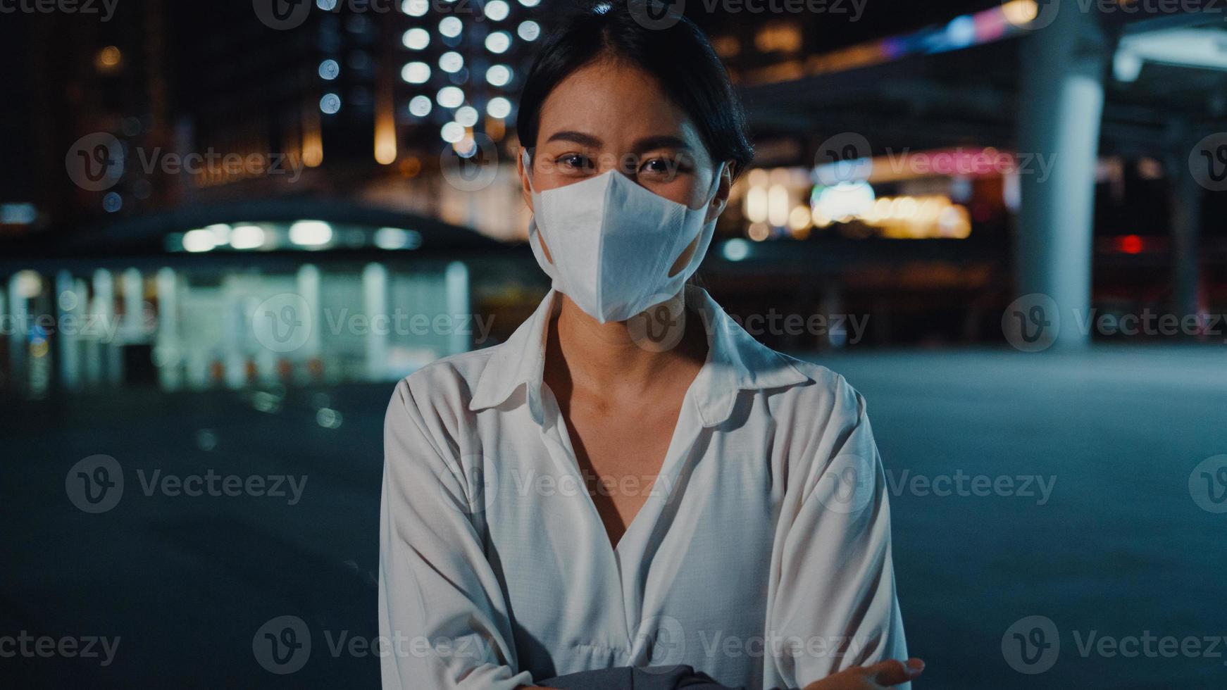 giovane donna d'affari di successo asiatica in abiti da ufficio di moda indossa una maschera medica sorridente e guardando la fotocamera mentre felice sta da sola all'aperto nella notte urbana moderna della città. concetto di affari in movimento. foto