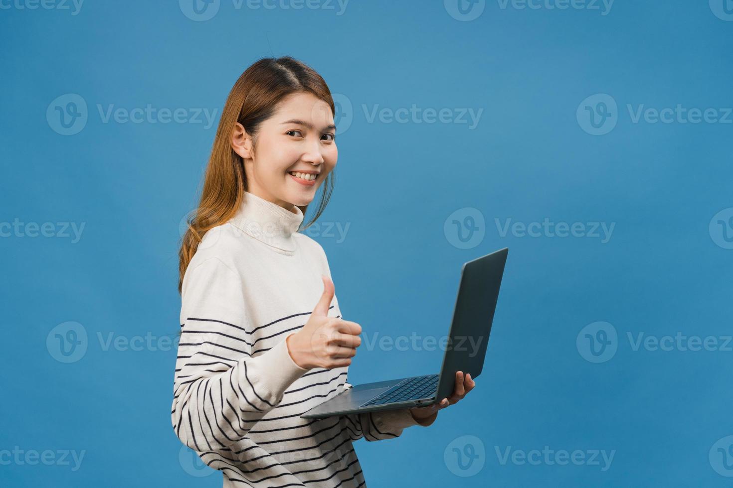 giovane donna asiatica che utilizza il computer portatile con espressione positiva, sorride ampiamente, vestita con abiti casual sentendosi felici e in piedi isolato su sfondo blu. felice adorabile donna felice esulta successo. foto