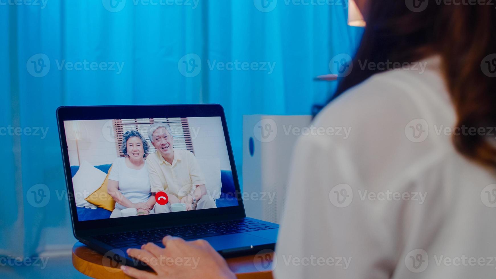 Scambio studentesco asiatico femmina utilizzando la videochiamata portatile parlando con la famiglia mentre si lavora da casa in soggiorno di notte. autoisolamento, distanza sociale, quarantena per coronavirus nella prossima normalità. foto