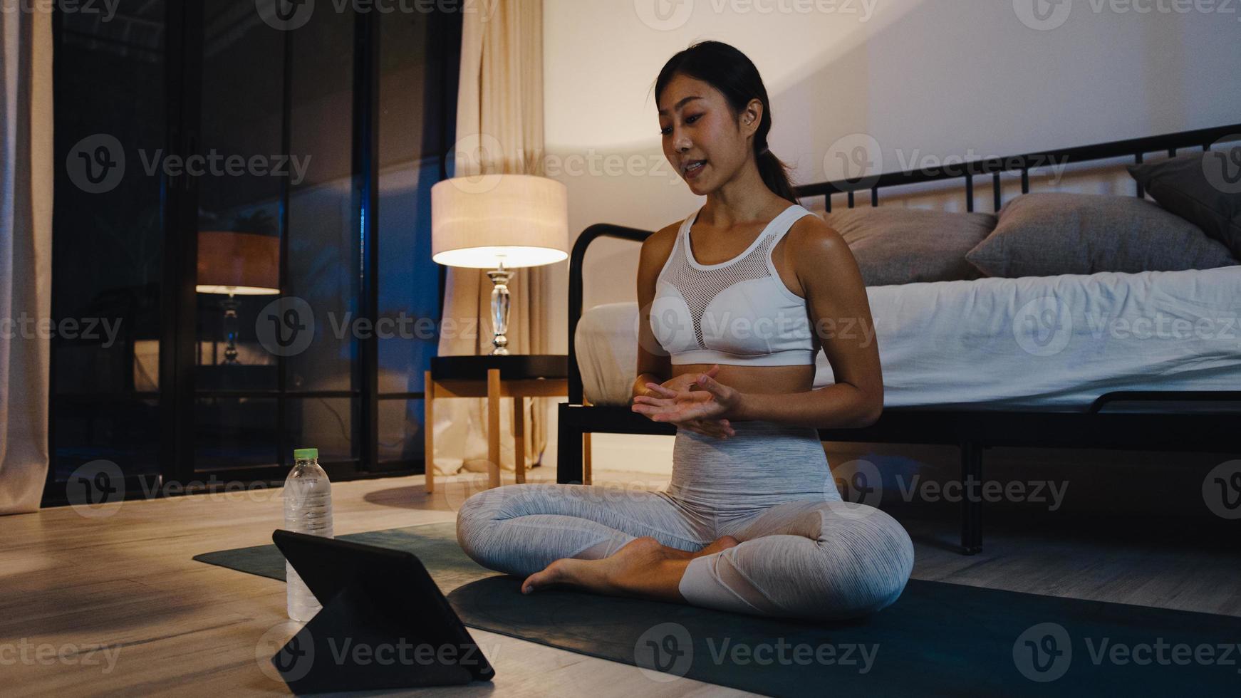 giovane donna asiatica in esercizi di abbigliamento sportivo facendo esercizio e utilizzando tablet per guardare video tutorial di yoga a casa la notte. formazione a distanza con personal trainer, distanza sociale, concetto di educazione online. foto