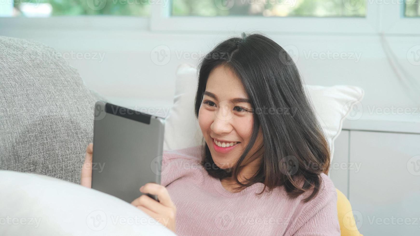 donna asiatica che ascolta musica e usa tablet, donna che usa il tempo di relax sdraiata sul divano di casa nel soggiorno di casa. musica d'ascolto femminile felice con il concetto delle cuffie. foto