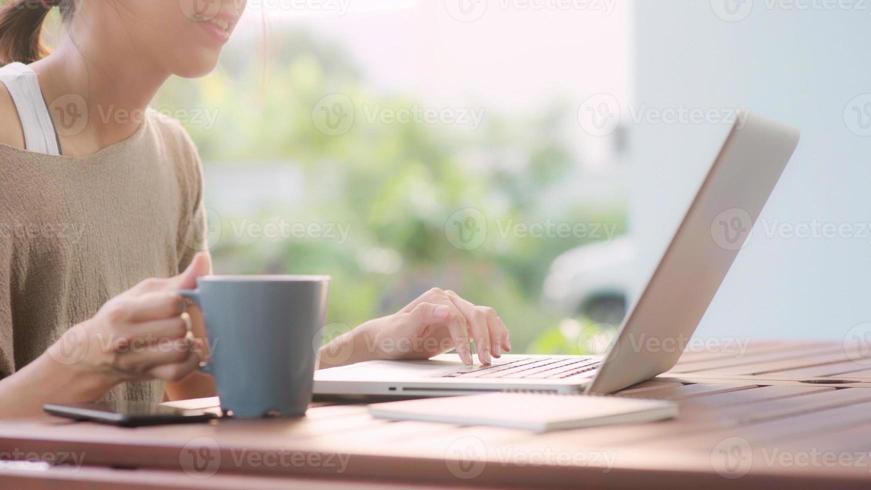 donna asiatica freelance che lavora a casa, donna d'affari che lavora al computer portatile e beve caffè seduta sul tavolo in giardino al mattino. donne di stile di vita che lavorano a casa concetto. foto