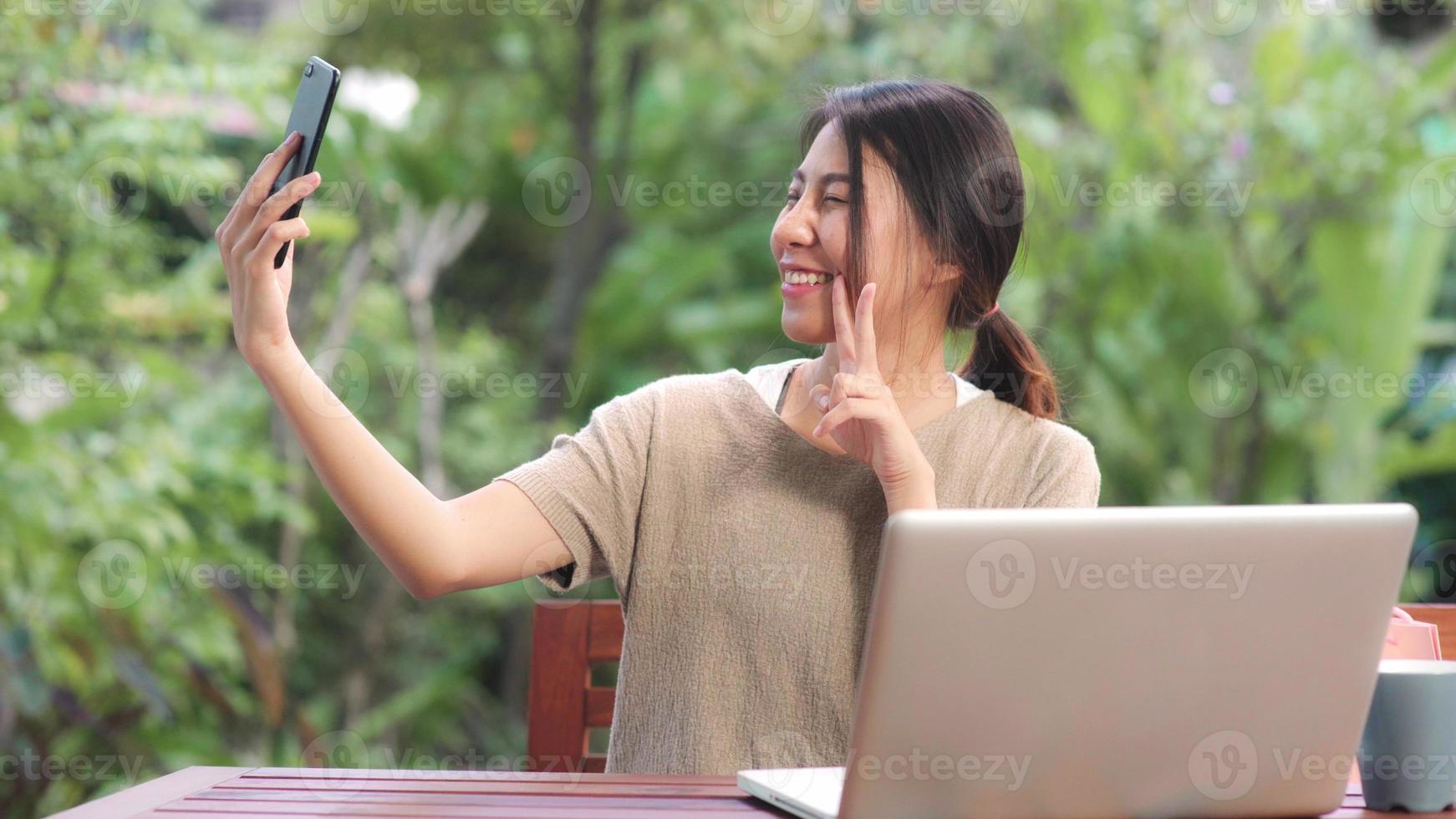 donna asiatica che utilizza il post selfie del telefono cellulare nei social media, la donna si rilassa sentendosi felice mostrando le borse della spesa seduta sul tavolo in giardino al mattino. le donne di stile di vita si rilassano a casa concetto. foto