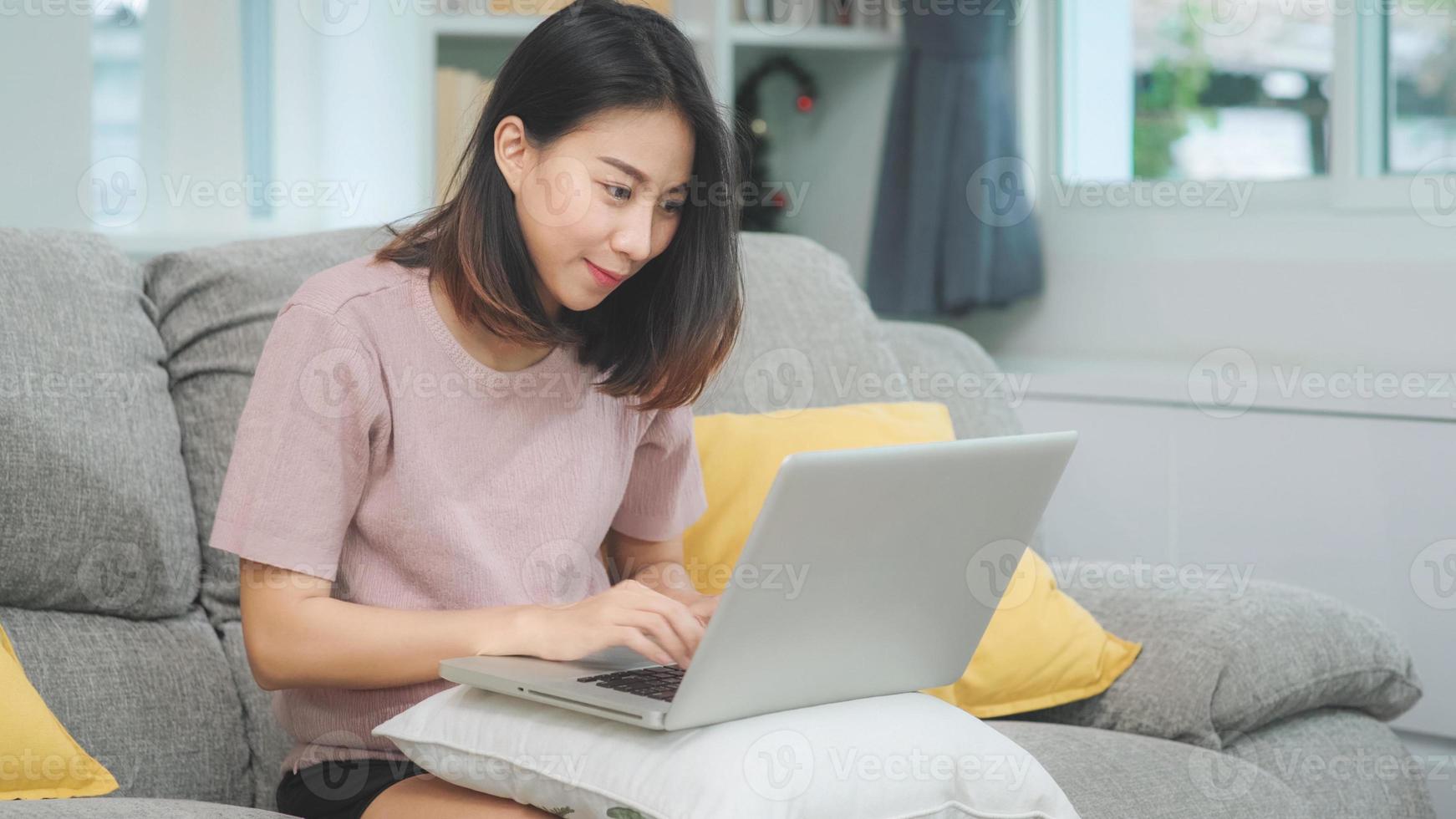 giovane donna asiatica freelance d'affari che lavora al computer portatile controllando i social media mentre è sdraiata sul divano quando si rilassa nel soggiorno di casa. stile di vita latino e donne ispaniche di etnia al concetto di casa. foto