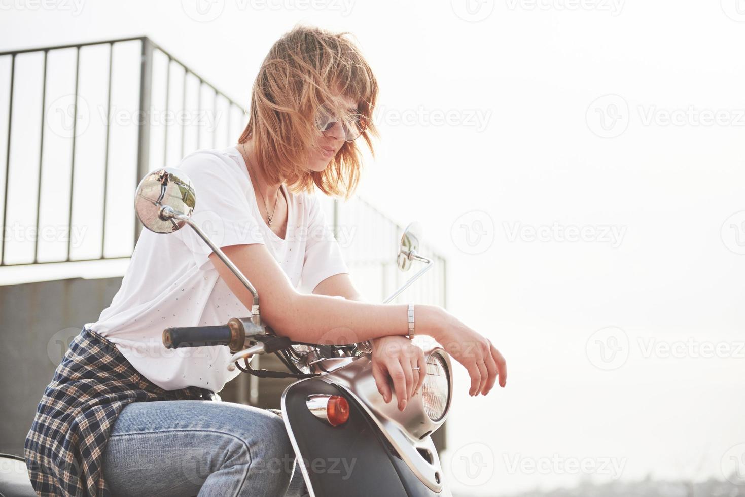 ritratto di una bella ragazza hipster seduta su uno scooter retrò nero, sorridente in posa e godersi il caldo sole primaverile. foto