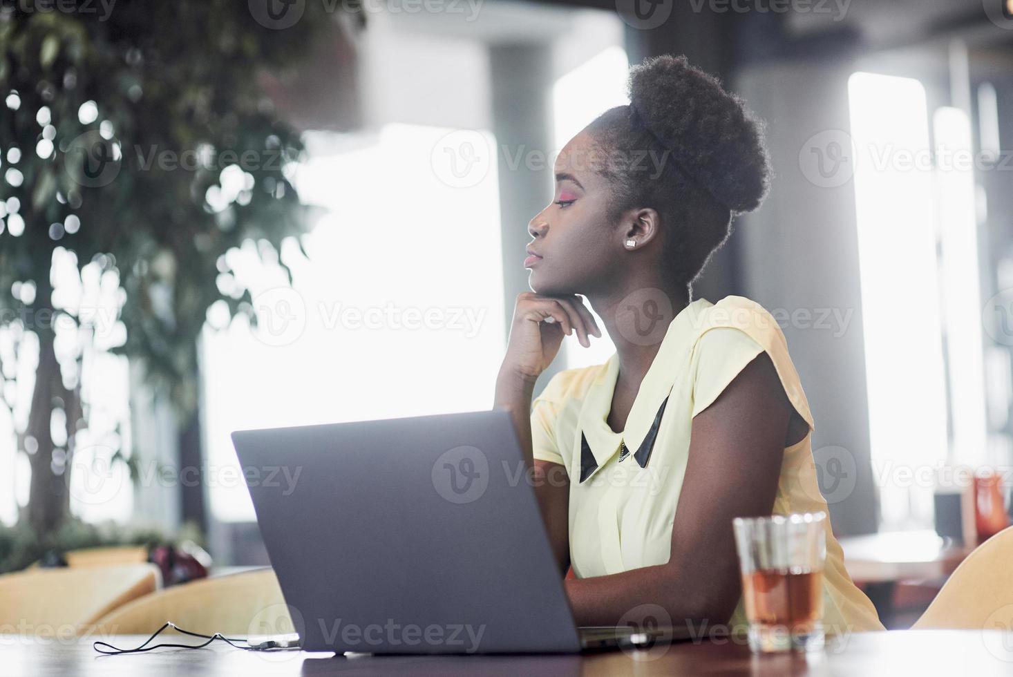 una giovane ragazza afroamericana con i capelli ricci scuri che riflette su un laptop in un bar foto