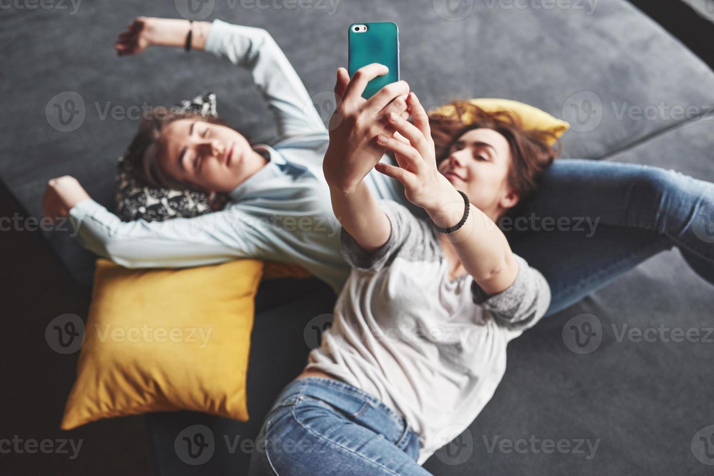 due sorelle gemelle sorridenti carine che tengono smartphone e fanno selfie. le ragazze si sdraiano sul divano in posa e gioia foto