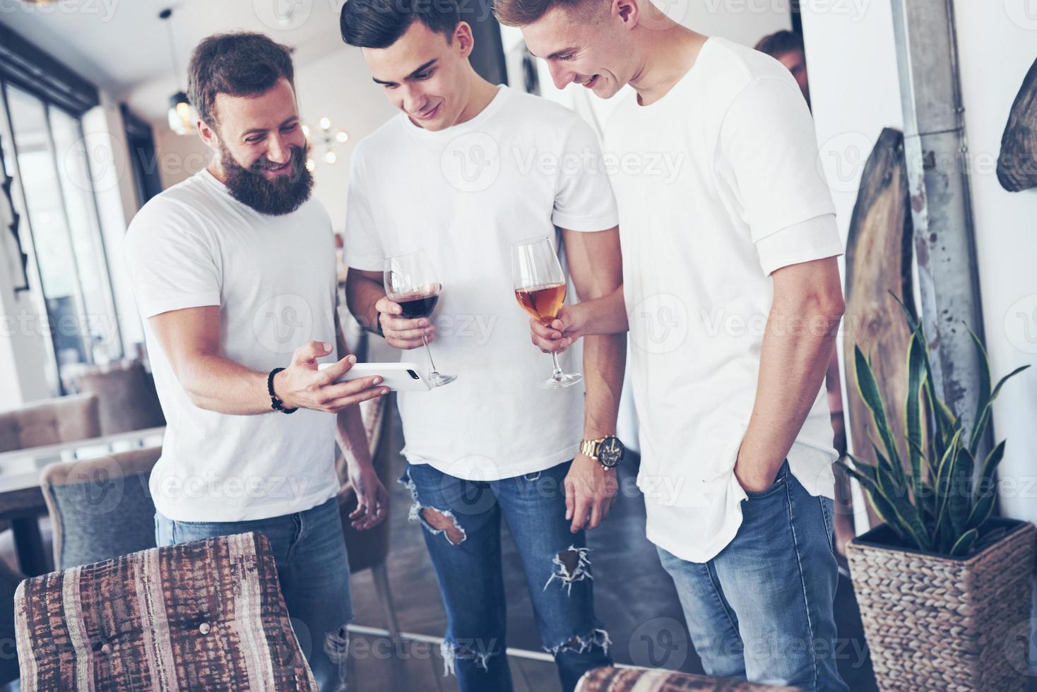 vecchi amici allegri comunicano tra loro e guardano il telefono, con bicchieri di whisky o vino in pub. concetto di intrattenimento e stile di vita. persone connesse wifi nella riunione del tavolo del bar foto