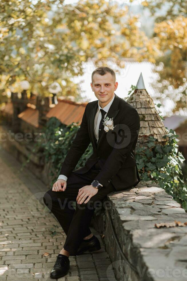 il giovane sposo si siede su il recinto guardare in il lente. un adulto uomo nel un' nero completo da uomo e bianca camicia con un' cravatta foto