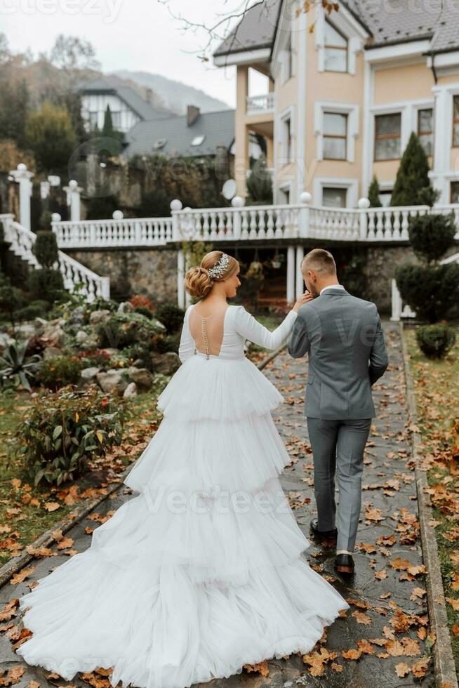 un' camminare di il sposa e sposo nel un' parco con bellissimo architettura, bianca ringhiere. sposa con un' lungo treno foto