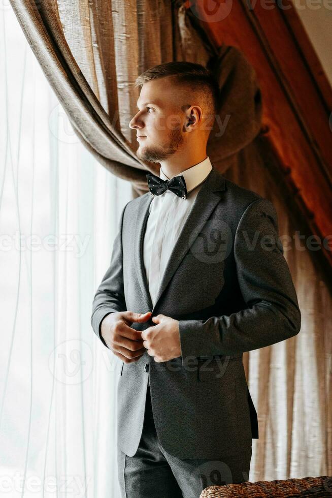 un' giovane uomo d'affari nel un' Hotel camera è vestito nel un' classico grigio completo da uomo, un' bianca camicia e un' arco legare. riposo nel meraviglioso appartamenti. quello dello sposo mattina a il Hotel. maschio mani vicino su. ritagliata foto. foto