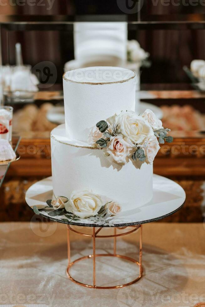 un' bianca a due livelli nozze torta, decorato con bianca fiori e cenere in giro il bordi, sta su un' In piedi vicino un' specchio. nozze dolci. foto