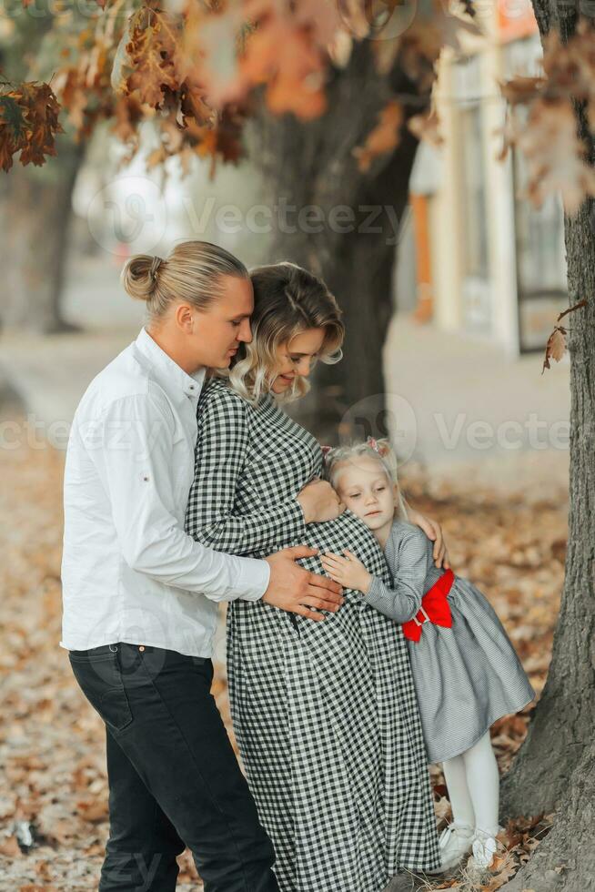 ritratto di un' giovane incinta donna con sua famiglia al di fuori nel un' parco tra ingiallito le foglie foto