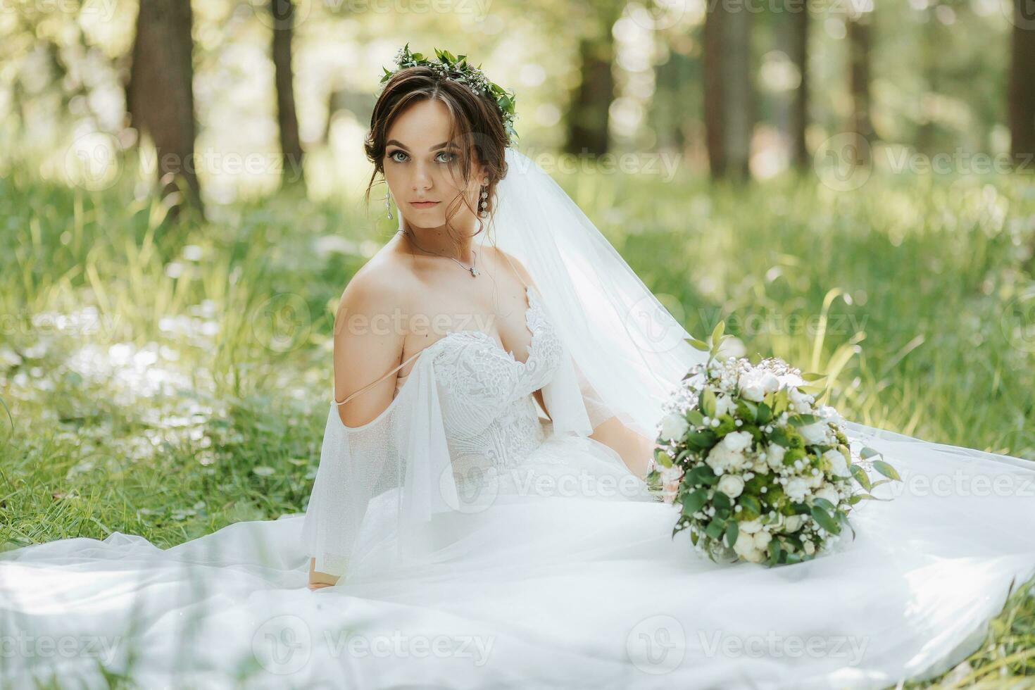 un' bellissimo sposa nel un' bianca vestito con un' ghirlanda di fresco fiori è seduta su il verde erba foto