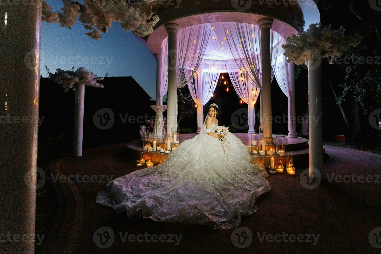 un' giovane bellissimo sposa nel un' nozze vestito con Aperto le spalle e un' corona su sua testa è seduta su il le scale nel il sera luci dopo il nozze cerimonia. foto