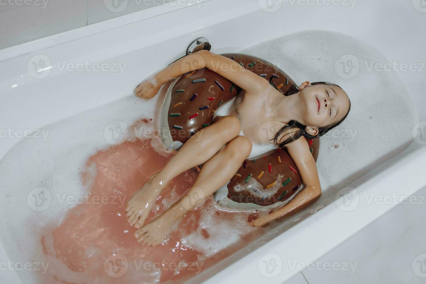 piccolo, sorridente, bellissimo dai capelli scuri ragazza con lungo capelli, bambino fa il bagno, lavaggi nel un' bianca bagno con schiuma. divertimento foto. foto